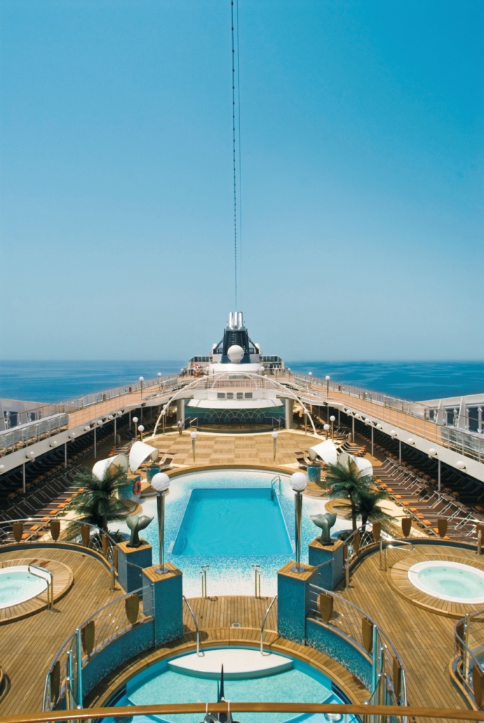 Blick auf das Deck eines Kreuzfahrtschiffs mit zwei Pools, Liegestühlen und dekorativen Palmen unter einem klaren blauen Himmel.