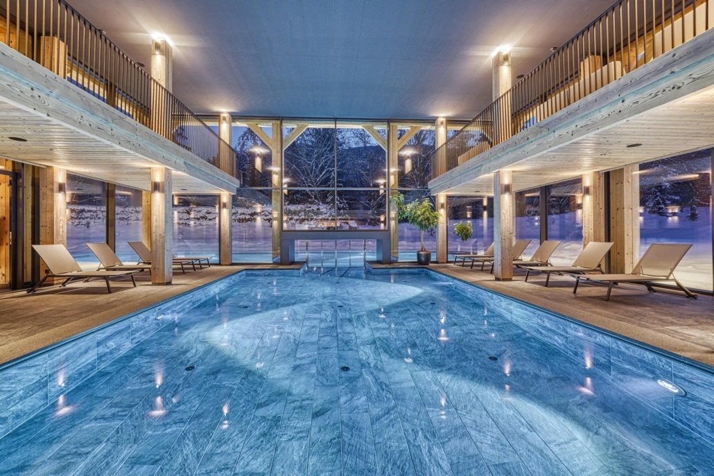 Innenpool mit Liegestühlen auf beiden Seiten, großen Fenstern mit Blick auf eine verschneite Landschaft und warmer Umgebungsbeleuchtung – perfekt zum Entspannen