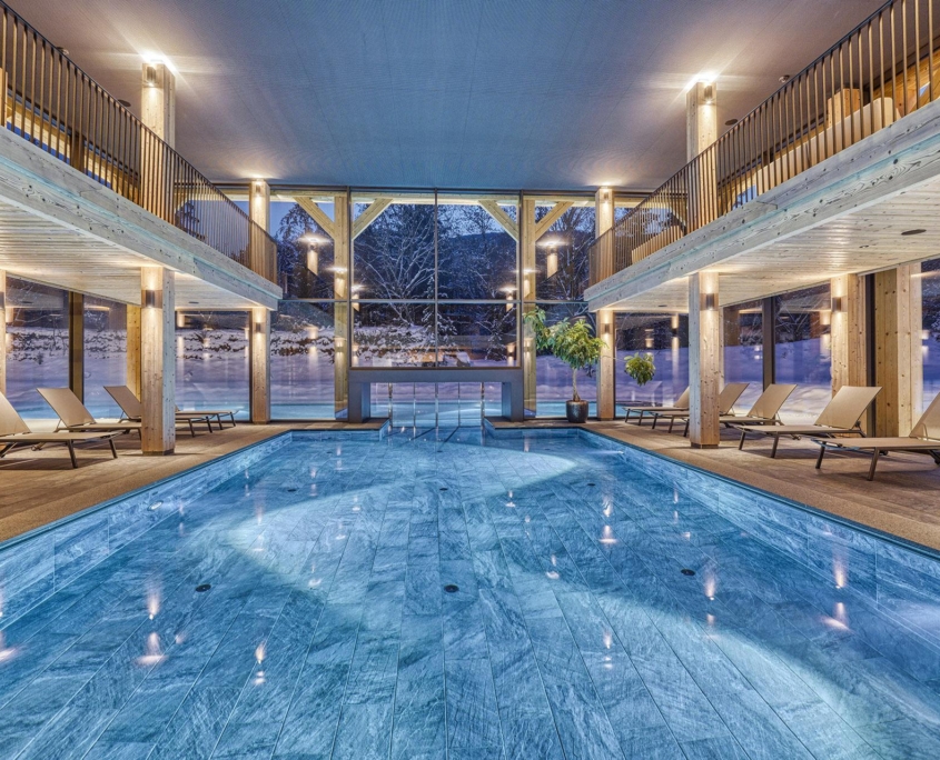 Innenpool mit Liegestühlen auf beiden Seiten, großen Fenstern mit Blick auf eine verschneite Landschaft und warmer Umgebungsbeleuchtung – perfekt zum Entspannen