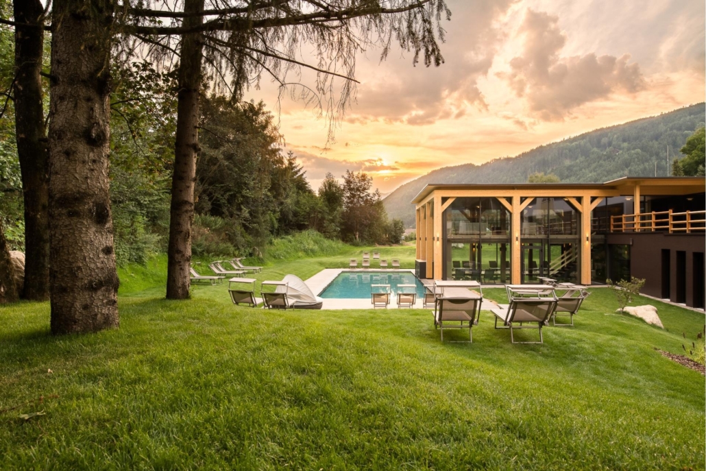 Eine Rasenfläche mit Liegestühlen umgibt einen Außenpool neben einem modernen Gebäude, vor der Kulisse von Bäumen und Bergen bei Sonnenuntergang – ein idealer Ort für eine Gruppenreise zum Lago Maggiore.