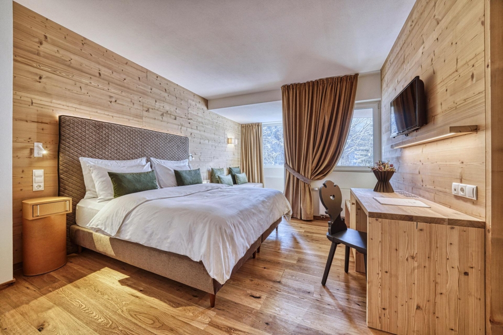 Ein modernes Hotelzimmer mit holzgetäfelten Wänden und Böden, einem großen Doppelbett, grünen Kissen, einem Schreibtisch mit Stuhl, einem an der Wand montierten Fernseher und einem Fenster mit braunen Vorhängen – perfekt zum Entspannen nach Ihrer Gruppenreise Lago Maggiore.