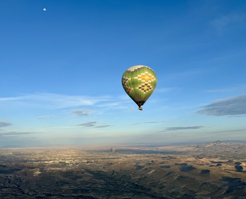 Ein grün-gelber Heißluftballon schwebt über einer weiten, sonnenbeschienenen Landschaft in der Nähe von Kappadokien, bei klarem Himmel und sichtbarem Mond.